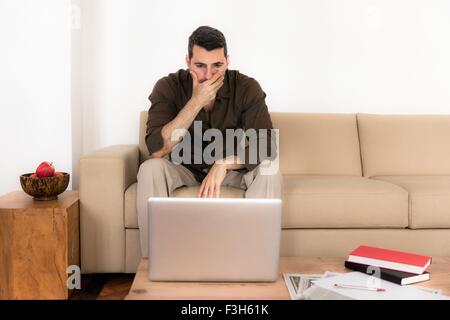 Mature man sitting on sofa looking at laptop, hand over mouth Stock Photo
