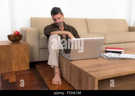 Barefoot mature man sitting on wooden floor with laptop, looking down Stock Photo