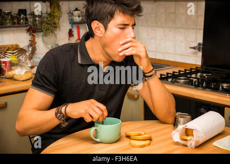 Tired Attractive Young Man Yawning with Eyes Closed and Hand on Mouth, Leaning with Elbow on Kitchen Table and Holding Cup of Co Stock Photo