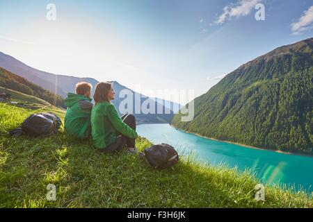 Young couple sitting  looking out over Vernagt reservoir and Finailhof farmhouse, Val Senales, South Tyrol, Italy Stock Photo