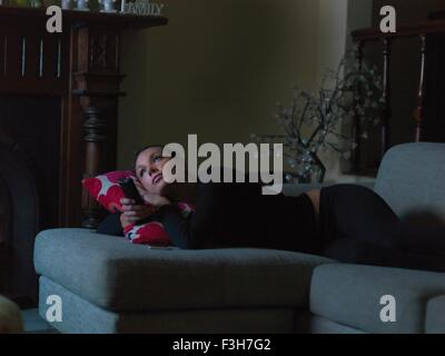 Mid adult woman lying on sofa, at night, watching TV, holding TV remote Stock Photo
