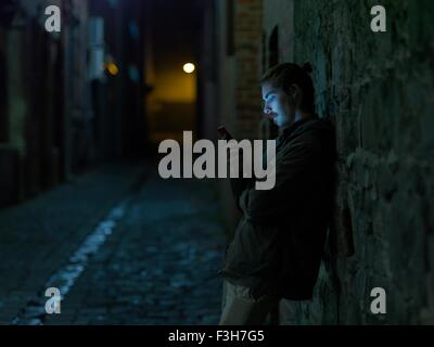 Young man in street, leaning against wall, at night, using smartphone Stock Photo