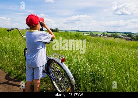 Young boy standing with bicycle beside field, looking at landscape through binoculars, rear view Stock Photo