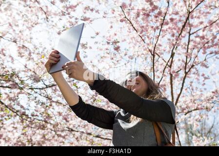 Mature woman taking digital tablet selfie in front of cherry blossom Stock Photo