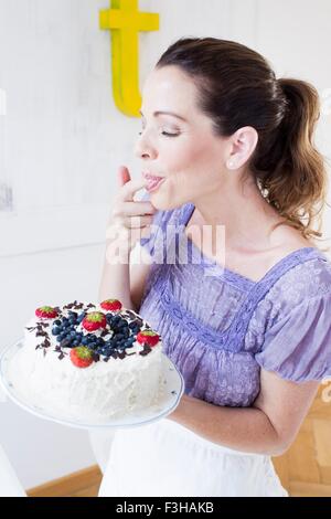 Mature woman holding fruit covered cake, licking fingers eyes closed Stock Photo