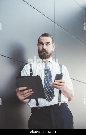 Stylish businessman using smartphone and digital tablet leaning against office wall Stock Photo
