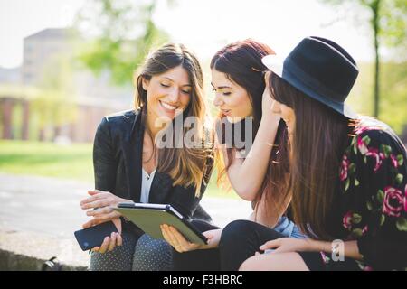 Three young female friends sharing update on digital tablet in park Stock Photo