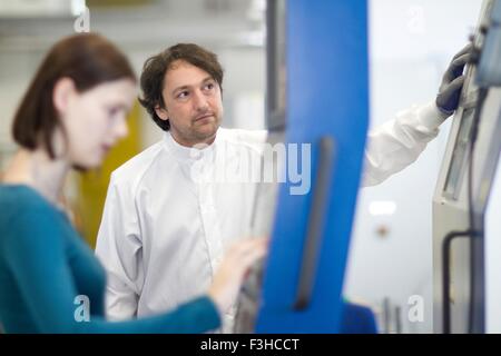 Mid adult man wearing labcoat and protective gloves operating machine, looking away Stock Photo