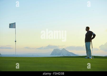 Golfer holding golf club standing near golf flag looking away Stock Photo