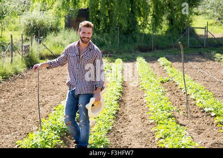 Young man holding hat standing in vegetable garden looking at camera smiling Stock Photo