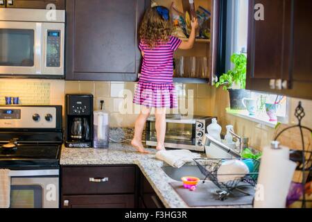 Young girl standing on kitchen work surface, looking for treats in cupboard, rear view Stock Photo