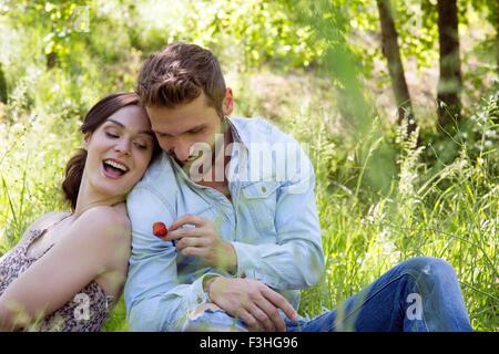 Young couple sitting back to back on grass holding strawberry smiling Stock Photo