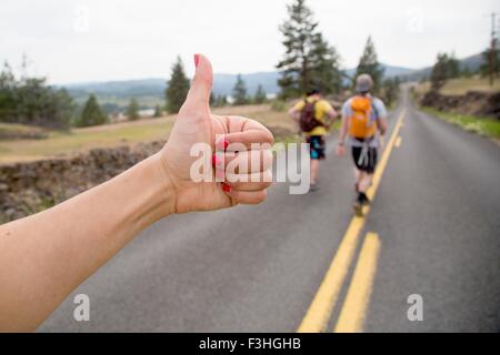 Two men hiking along road, while woman thumbs a lift, focus on hand Stock Photo