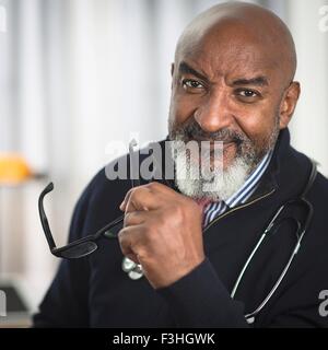 Portrait of mature doctor with stethoscope, holding glasses, looking at camera smiling Stock Photo