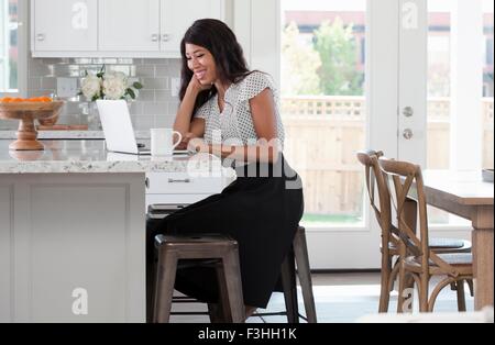 Young woman, sitting at kitchen counter with coffee and laptop Stock Photo