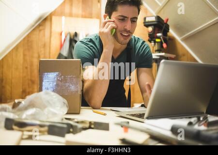 Young man in workshop sitting at desk talking on telephone looking at laptop Stock Photo