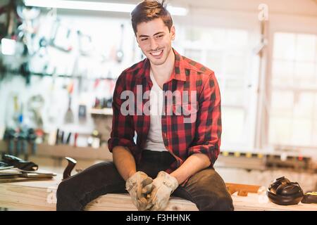 Young man sitting on workbench wearing protective gloves, looking away smiling Stock Photo