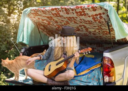 Young woman petting cat and playing ukulele whilst camping in pick up boot Stock Photo