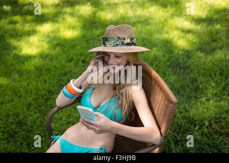 Young woman wearing bikini and sunhat reading smartphone texts in garden Stock Photo