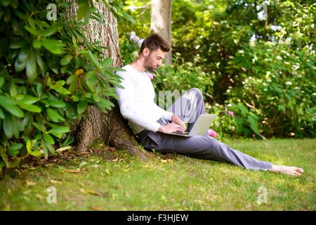Young man sitting against tree, using laptop Stock Photo