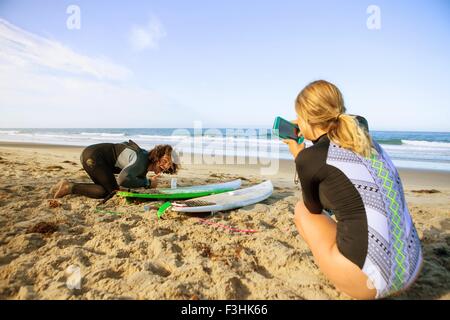 Couple on beach, young woman taking photograph of man with surfboard Stock Photo