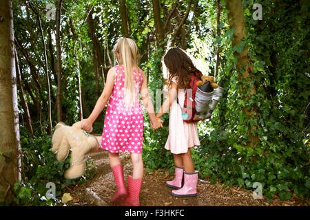 Rear view of two girls carrying teddy bear and backpack walking in forest Stock Photo