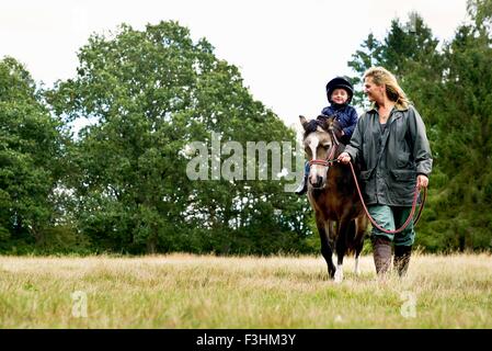 Mother leading horse riding son in field Stock Photo