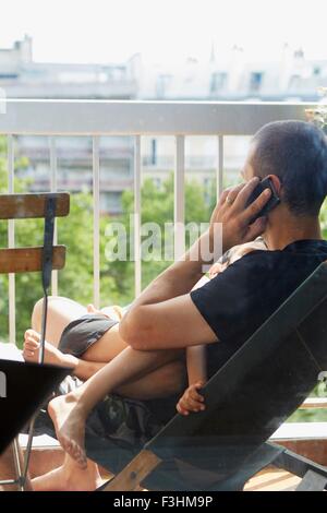 Father using smartphone and cradling sleeping son on balcony Stock Photo