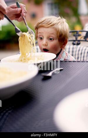 Boy sitting at garden table being served spaghetti, looking surprised Stock Photo
