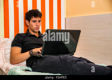 Attractive Young Man with Serious Expression, Reclining Comfortably with Laptop on Bed Working on his Start-up Business - Young Stock Photo