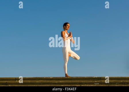 Young woman, wearing a white body suit, is practising Hatha-Yoga outdoor, showing the pose: vrikshasana, tree Stock Photo