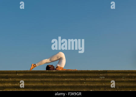 Young woman, wearing a white body suit, is practising Hatha-Yoga outdoor, showing the pose: halasana, plough Stock Photo