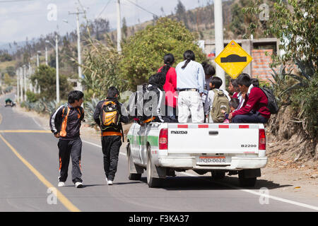 A group of students, some wearing school uniforms and carrying backpacks, are riding in the open bed of a white Mazda B2200 pickup truck on a paved rural road. Two other students in matching black Stock Photo
