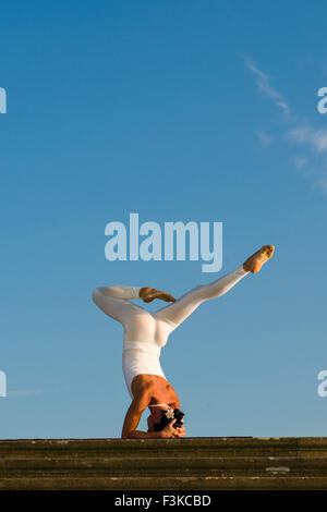 Young woman, wearing a white body suit, is practising Hatha-Yoga outdoor, showing the pose: sirshasana, head stand Stock Photo