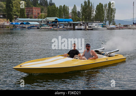 Two men in a yellow speedboat going slow through a marina with boats and buildings in the background. Stock Photo