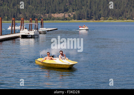 Two men in a yellow speedboat going slow through a marina with boats and a dock in the background. Stock Photo