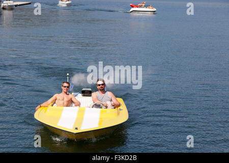 Two men in a yellow speedboat going slow through a marina with boats and a dock in the background. Stock Photo