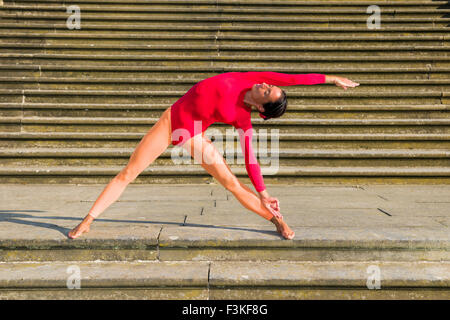 Young woman, wearing a red-orange body suit, is practising Hatha-Yoga outdoor, showing the pose: trikonasana, triangle Stock Photo