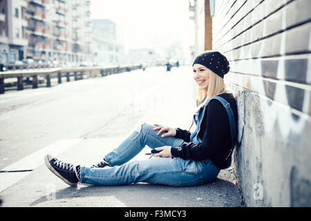 young beautiful blonde straight hair woman in the city, sitting on the ground, overlooking right, holding a smartphone, laughing - happiness, communication, technology concept Stock Photo