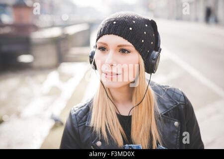 Portrait of young beautiful blonde straight hair woman in the city with headphones listening to music, overlooking right, smiling - happiness, relaxing concept Stock Photo