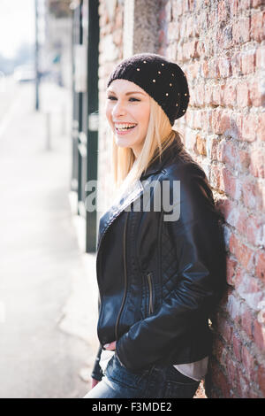 Knee figure of young beautiful blonde straight hair woman in the city leaning on a brick wall, overlooking left, laughing - happiness concept - wearing back leather jacket, jeans, white shirt Stock Photo
