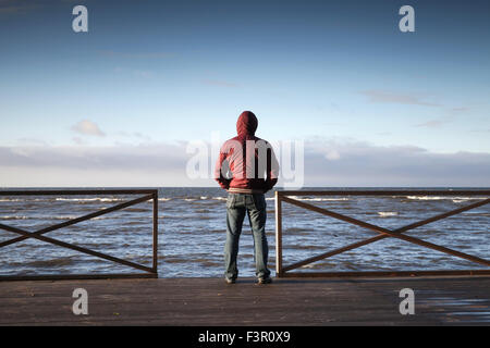 Young man in hood looking on the sea from wooden pier at morning. Rear view photo Stock Photo