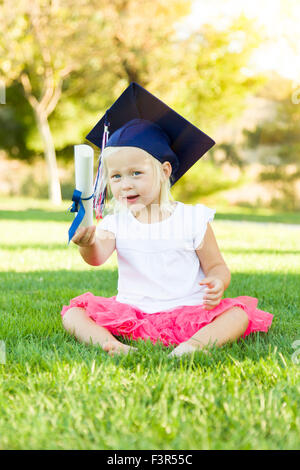 Cute Little Girl In Grass Wearing Graduation Cap Holding Diploma With Ribbon. Stock Photo