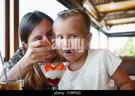 Mother feeds his daughter delicious sweet ice cream with strawberries in a restaurant. Shallow depth of field. Focus on the chil Stock Photo