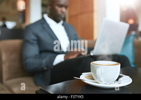 Refreshing hot cup of coffee on table with a businessman sitting in background working on laptop at hotel lobby. Stock Photo