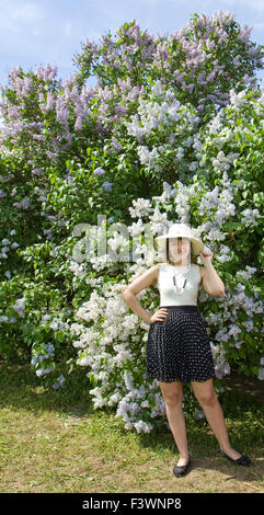 Lady, European, in hat and summer dress standing near big shrub of lilac in blossom. Stock Photo