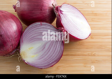 A large red onion that has been sliced in half on a wood cutting board with two additional onions to the side Stock Photo