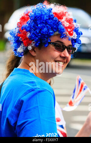 Man with red, white and blue hair, standing on side of street, wearing sunglasses and holding small Union jack flag. Turned towards viewer, smiling. Stock Photo