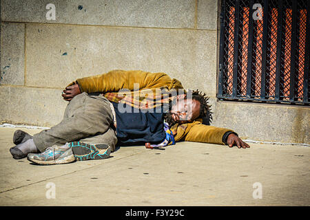 A dirty and homeless African American man sadly sleeps on a sidewalk on a city street in the USA Stock Photo