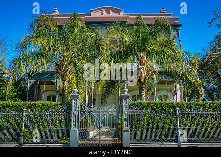 A decorative wrought iron fence & gate surrounds an historical mansion shrouded by palm trees in the Garden District in New Orleans, LA Stock Photo